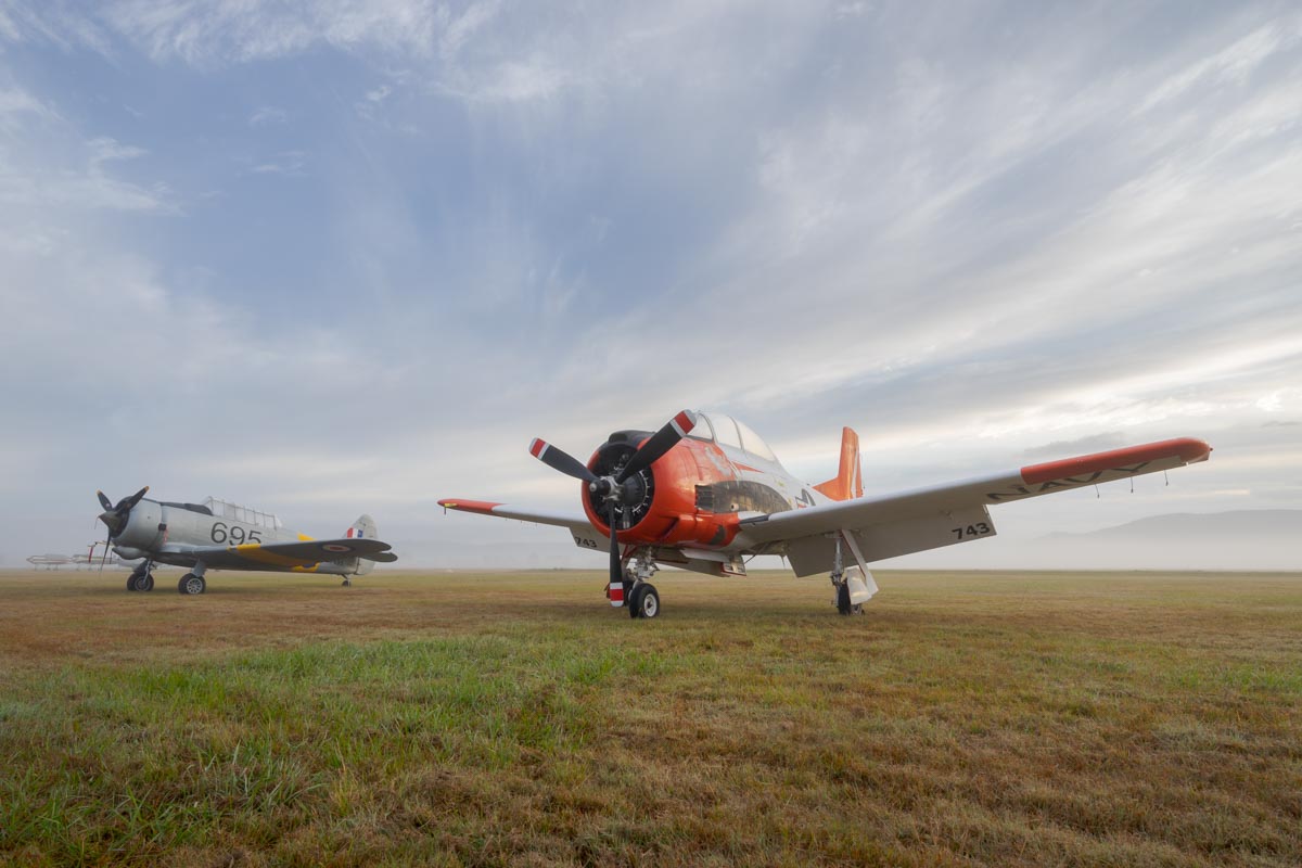 CAC CA-16 Wirraway VH-MFW and North American T-28B Trojan VH-RPX "Miss Stress" on the flight line in a foggy sunrise.